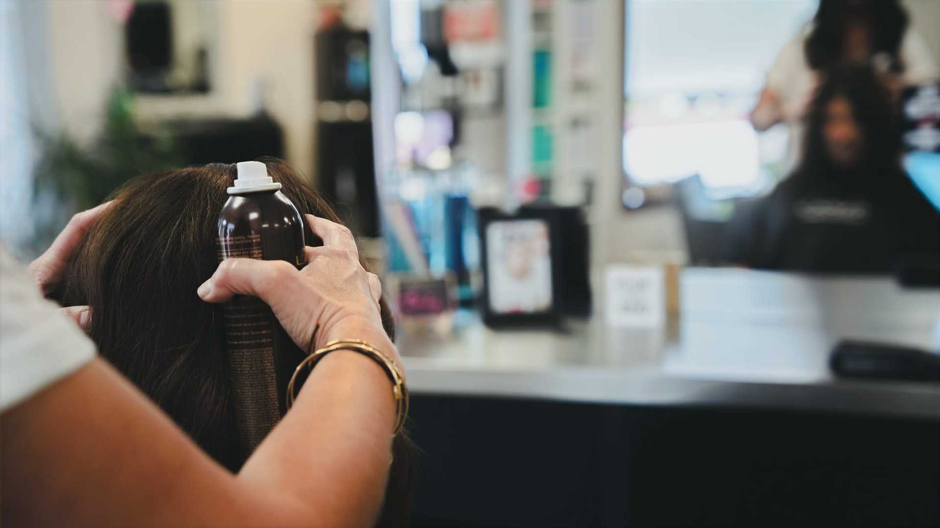 Hairdresser applies styling product to client's hair in a salon, with mirror in the background.